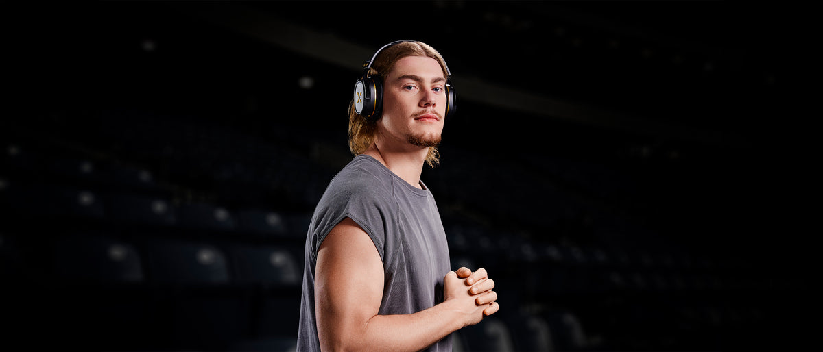 BlueAnt ambassador Harley Reid in a darkened stadium, wearing black and gold Pump X over-ear headphones.