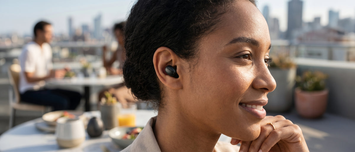 Smiling woman in her thirties wearing a black BlueAnt Pump Air Micro earbud at a rooftop café, city skyline behind