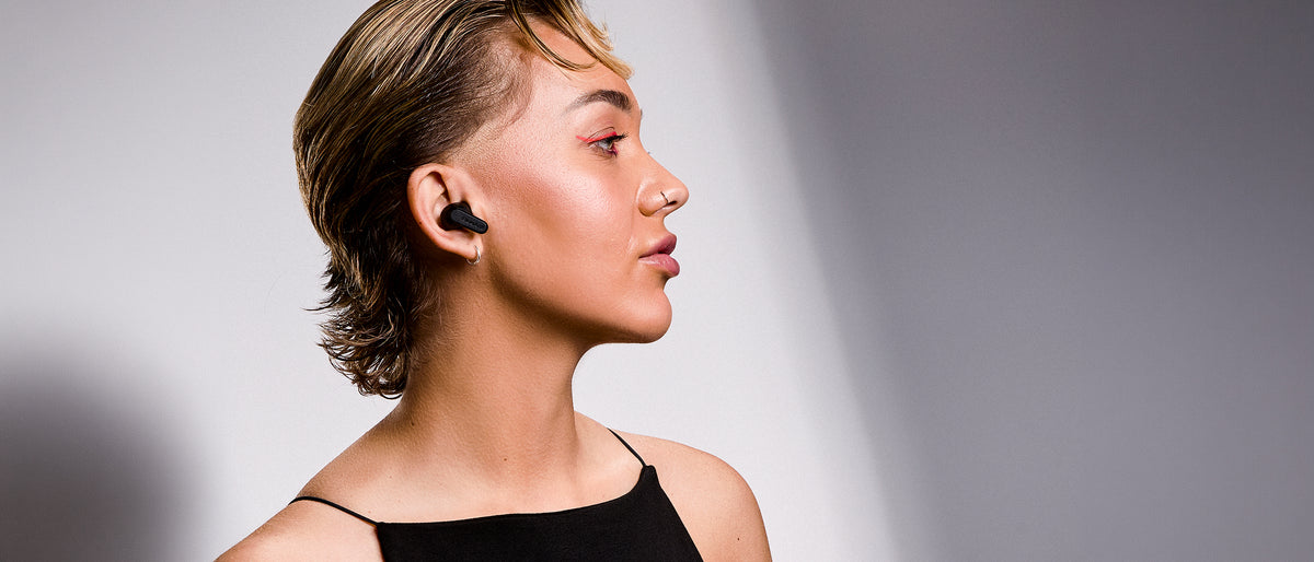 Woman wearing a black BlueAnt Pump Air Ultra earbud, side profile against a grey studio backdrop