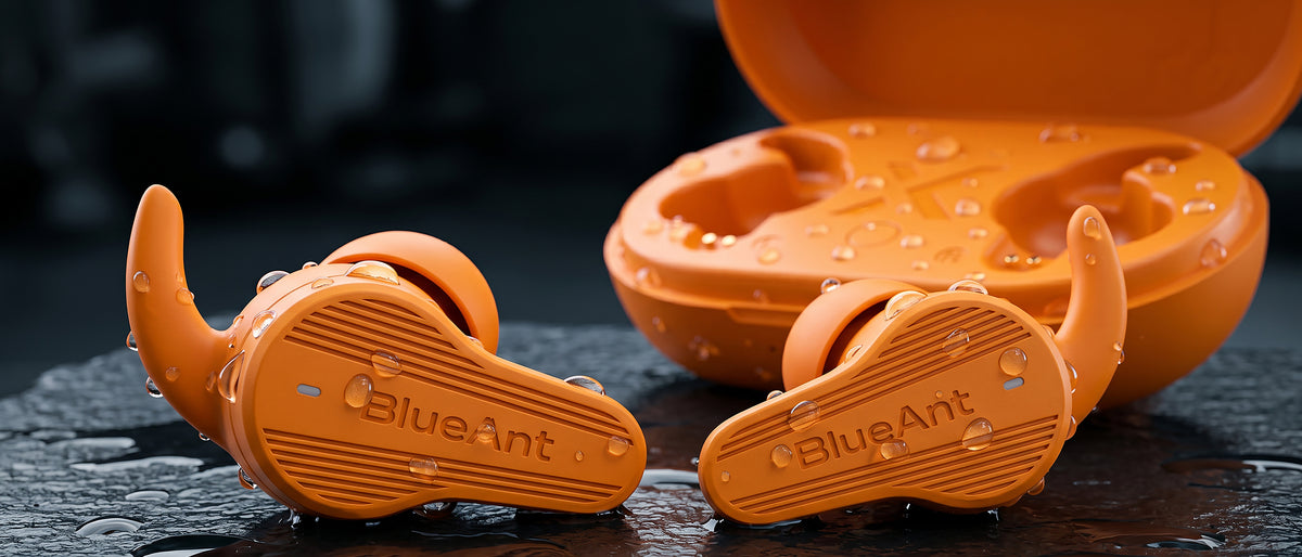 BlueAnt Pump Air X3 sport earbuds in orange with wing tips and open case, wet with water droplets