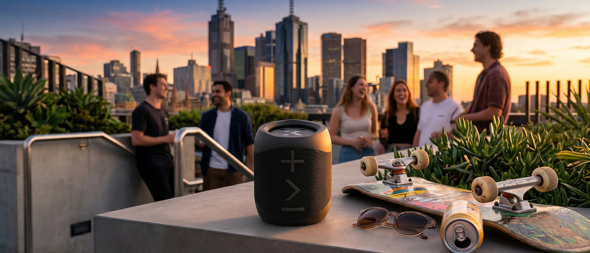 BlueAnt X1i speaker beside a skateboard and sunglasses at a rooftop gathering, friends laughing behind