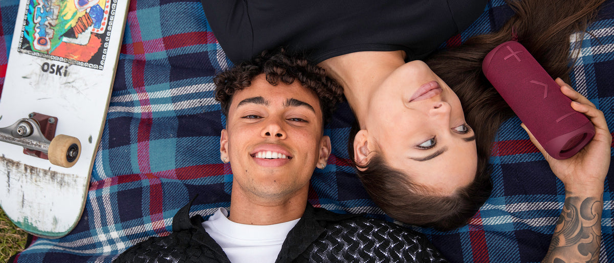 Two friends lying on a tartan picnic rug, one holding a maroon X2i speaker.