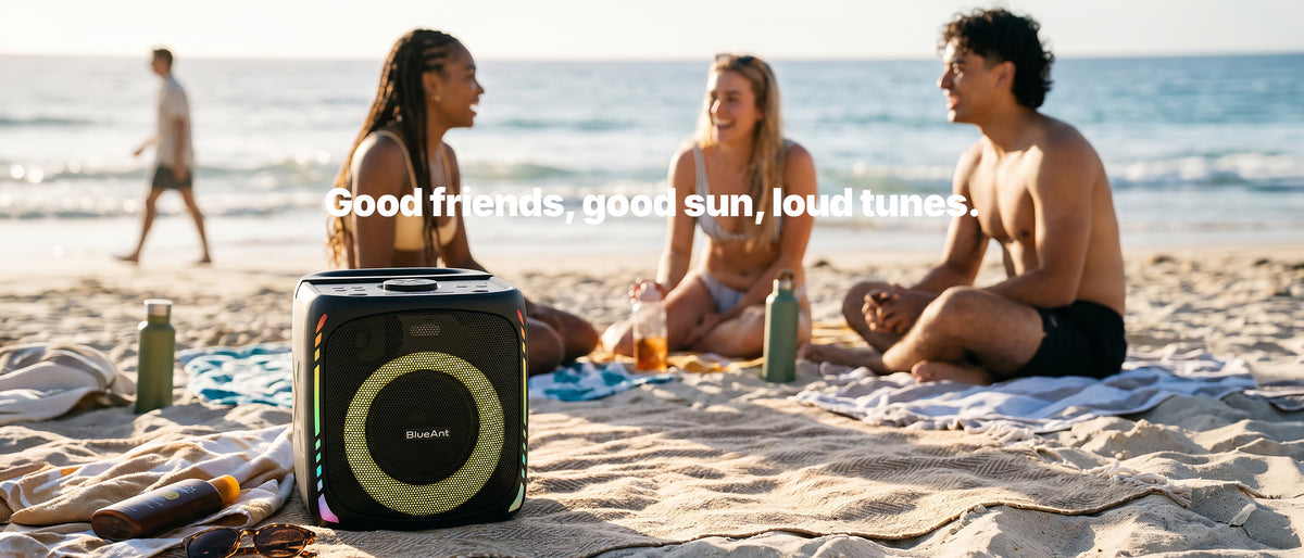 BlueAnt X4i portable party speaker with glowing RGB light ring on a beach towel, as three friends laugh on the sand by the ocean